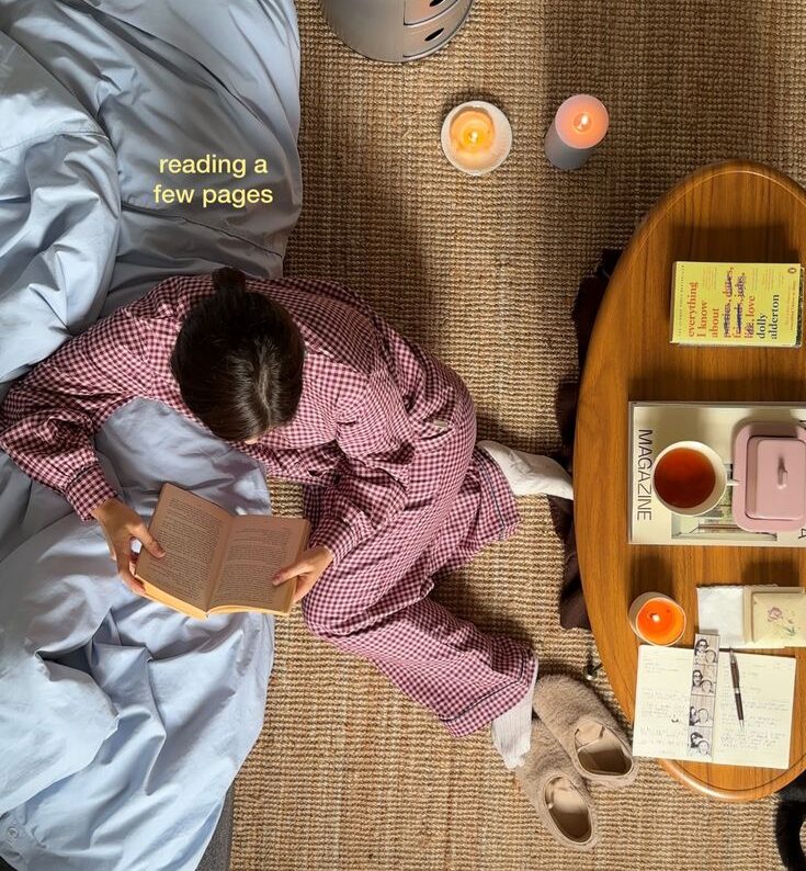 "Overhead view of a woman in checkered pajamas reading on the floor with tea and candles"